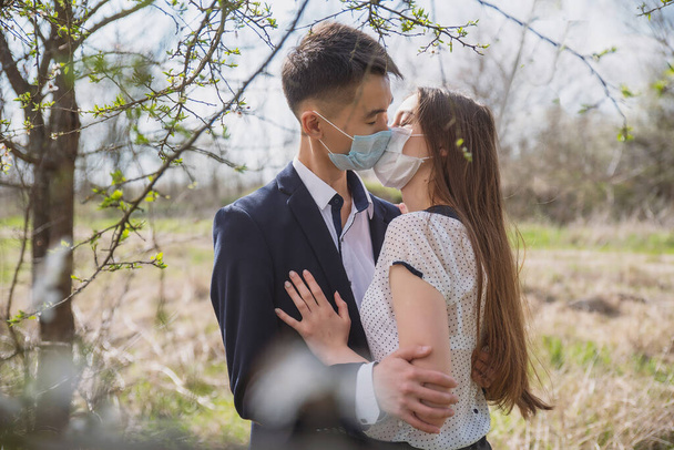 Asian guy with a European girl in medical masks on the background of flowering trees hug and kiss - Valokuva, kuva