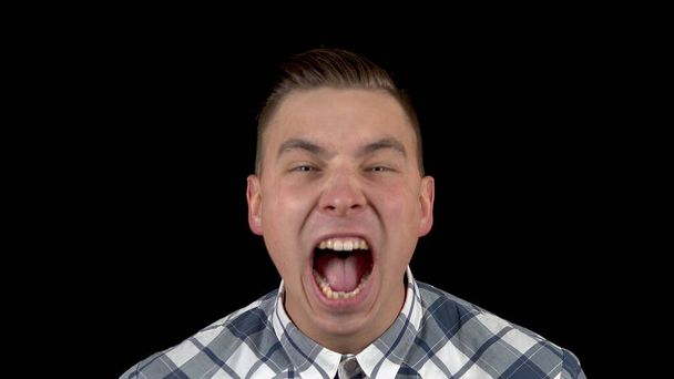 The young man is screaming. A man screams strongly in a shirt on a black background closeup - Photo, Image