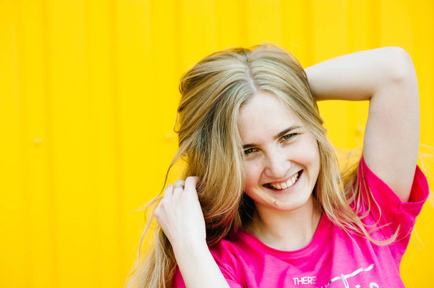 Beautiful athletic young woman with long blond hair in a pink shirt. Posing and smiling at wall of garage on yellow background. Place for text. close-up. Pretty white, straight teeth. Happy smile - Zdjęcie, obraz