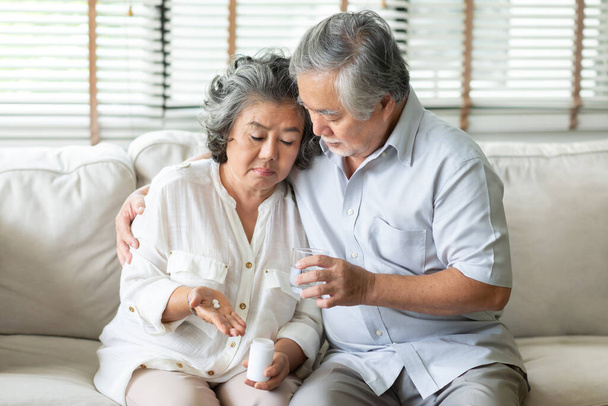 Starý muž utěšuje svou ženu, když je nachlazená. Starší žena drží pilulky nebo léky na ruce, zatímco má horečku a sedí doma na pohovce. Nemoc, Nemoc, Koronavirus, Virus, Covid-19, Karanténa. - Fotografie, Obrázek
