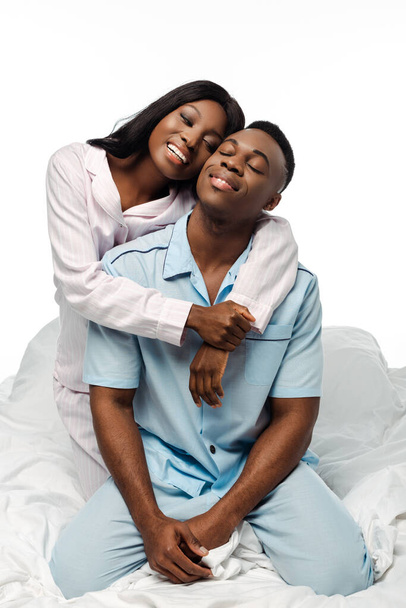ευτυχισμένο ζευγάρι Αφροαμερικανών που αγκαλιάζονται στο κρεβάτι με πιτζάμες απομονωμένες στα λευκά - Φωτογραφία, εικόνα
