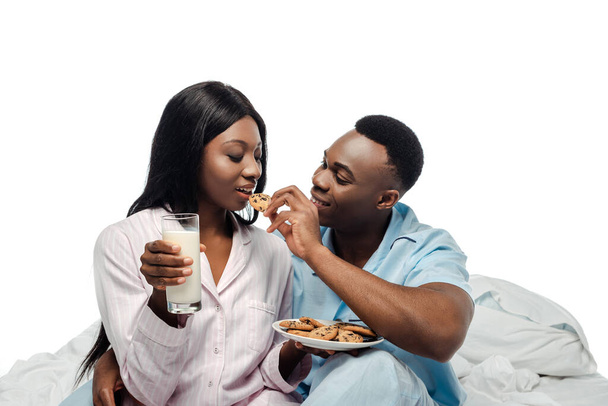 happy african american man feeding girlfriend with cookies in bed in pajamas isolated on white - Фото, изображение