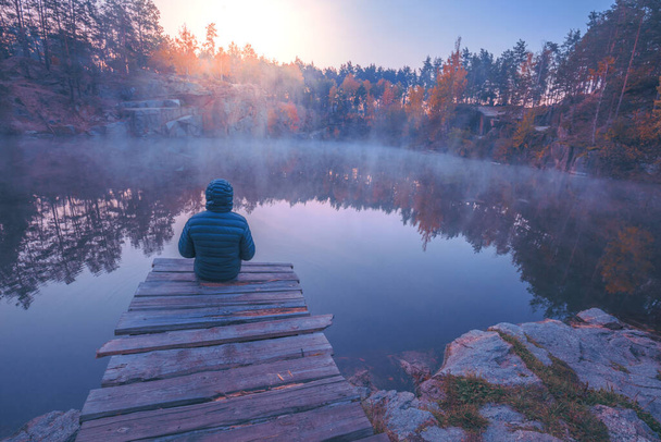 Un hombre sentado en una cubierta de madera y mirando a un lago con una orilla de granito. Salida del sol sobre el lago
 - Foto, Imagen