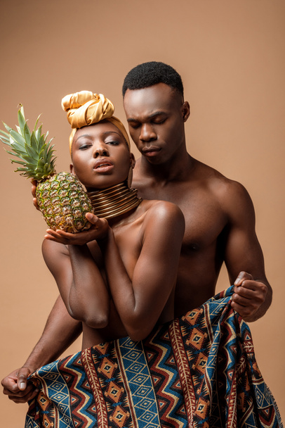 sexy naked tribal afro woman covered in blanket posing with pineapple near man on beige - Photo, Image