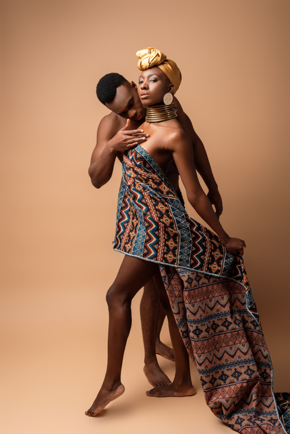 сексуальная обнаженная женщина племени афро, покрытая одеялом, позирующая рядом с мужчиной на бежевом
 - Фото, изображение