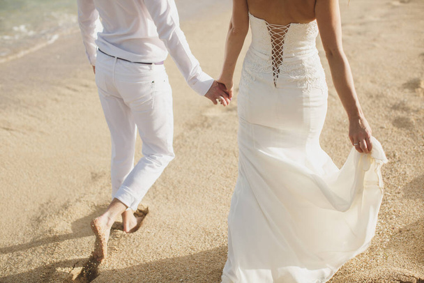 Η νύφη και ο γαμπρός περπατούν χέρι-χέρι στην άμμο. πατημασιές στην άμμο κοντά στον ωκεανό. - Φωτογραφία, εικόνα