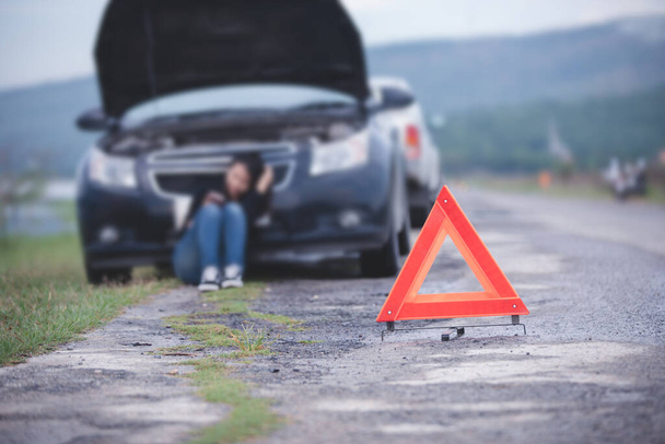 Mujer asiática sentada en el suelo cerca de coche roto con el triángulo rojo de un coche en la carretera
  - Foto, imagen
