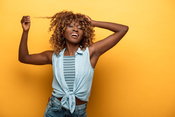 happy african american woman touching hair on yellow background - Foto, imagen