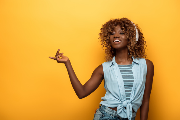 heureuse femme afro-américaine écouter de la musique dans les écouteurs et pointant du doigt sur fond jaune
 - Photo, image