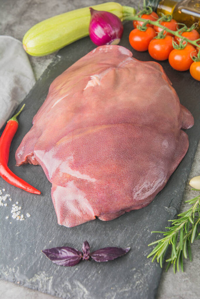 A large piece of raw pork liver on black slate with seasonings and spices in a butcher's shop, side view - Photo, Image