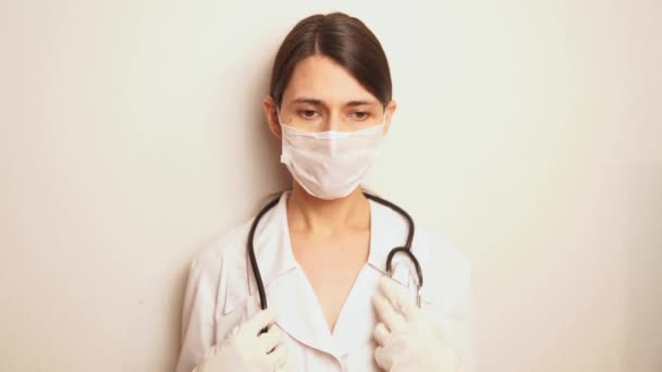 μια γυναίκα γιατρός σε ένα λευκό μιας χρήσης προστατευτική μάσκα και γάντια μιας χρήσης δείχνει εντάξει με το δεξί της χέρι τότε με το αριστερό της χέρι, γνέφει το κεφάλι της, είναι σίγουρος ότι όλα θα πάνε καλά - Πλάνα, βίντεο