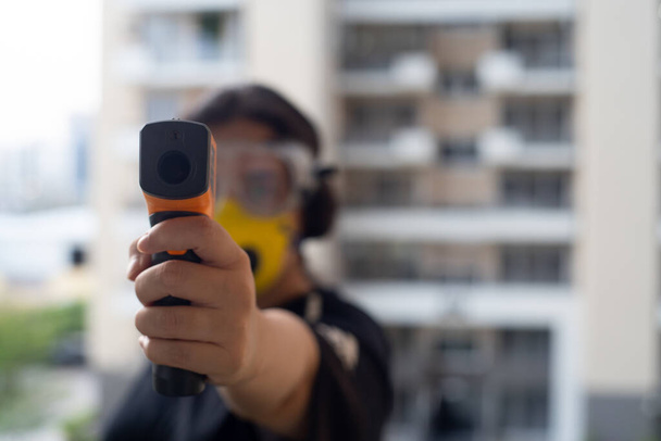 Young indian girl holding an infrared non contact thermometer to check the temperature with out of focus office residential background - Φωτογραφία, εικόνα