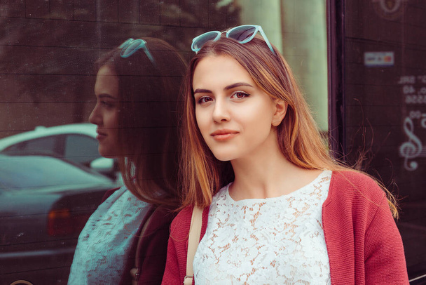 The girl stands by the window of the store office, smiles and looks at the camera, you outside outdoors in town city - Фото, изображение