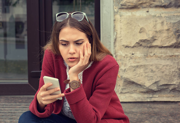 Woman preoccupied looking at her phone sitting near her house apartment building outdoors outside background - Foto, Bild