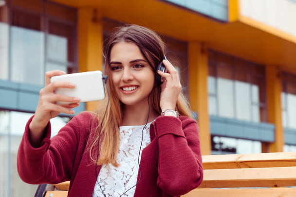 Pretty young woman using smartphone in headphones while sitting on bench.  - Foto, Imagen