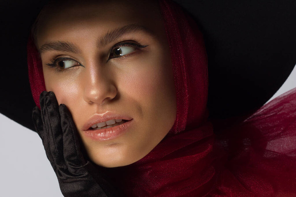 Fashionable beauty of a woman with a hat, black eyeliner and a red scarf. Clean and shine skin, natural lip gloss, thick eyebrows and long eyelashes. Vintage photography, cosmetology and makeup. - Photo, Image