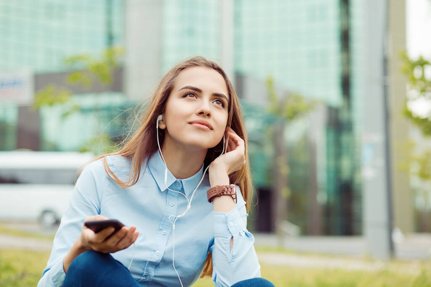 Listening to music audio book. Closeup business woman blue shirt sitting on grass hand on earphones having a conversation on phone looking up thinking how to solve the problem outdoors outside in city - Фото, зображення