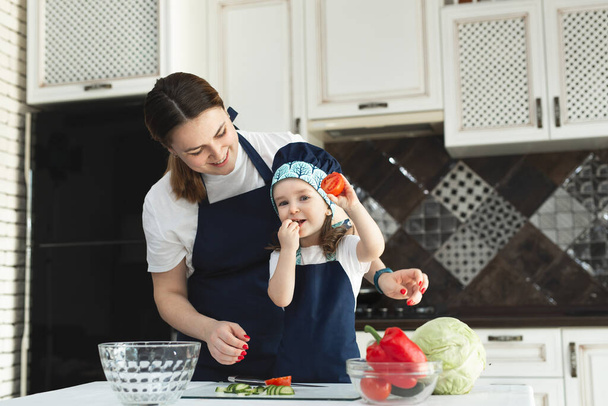幼い娘に台所でサラダを調理することを教える、若いお母さんと可愛い女の子子供はエプロンを身に着けていますまな板の上にナイフで野菜をみじん切り,自宅でキッチンに立って. - 写真・画像