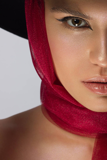 Half face beauty of a woman with a hat, black eyeliner and a red scarf. Clean and shine skin, natural lip gloss, thick eyebrows and long eyelashes. Vintage photography, cosmetology and makeup. - Foto, Bild