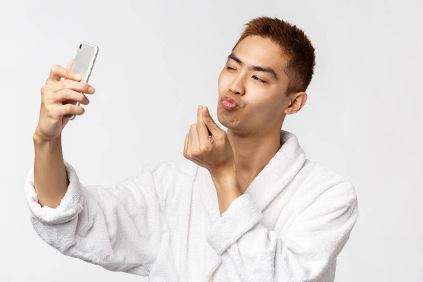 Концепция красоты, красоты и технологий. Портрет наглого молодого красивого азиатского мужчину в халате, лицо целующегося перед мобильной камерой, смартфон с селфи, белый фон
 - Фото, изображение