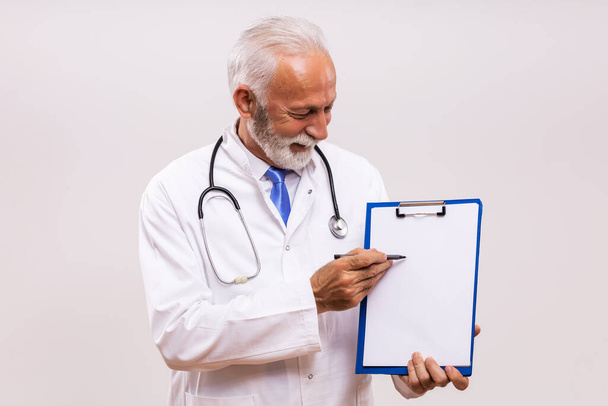 Portrait of senior doctor showing with pen clipboard on gray background. - Фото, изображение