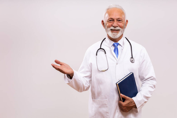 Portrait of senior doctor holding personal organizer and  showing welcome gesture on gray background. - Foto, immagini