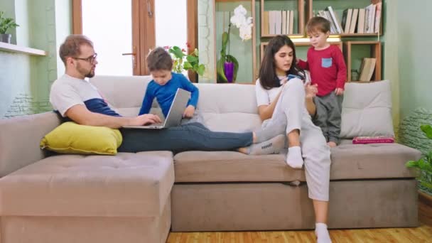 Impressionante jovem família com atraente dois filhos têm tempo de família juntos eles conversando brincando no sofá pai trabalhando em seu laptop pela maneira como todos estão felizes
 - Filmagem, Vídeo