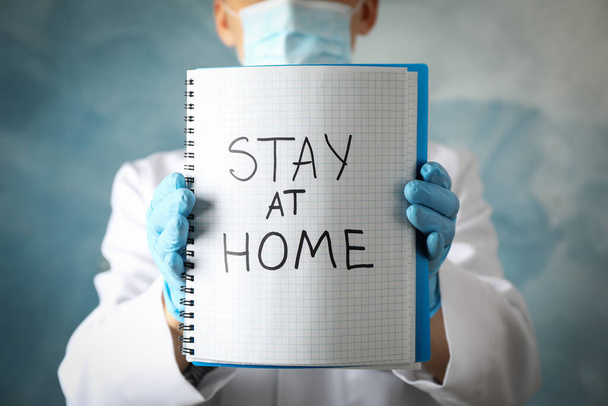 El médico sostiene el cuaderno con la inscripción Quédese en casa sobre fondo azul
 - Foto, Imagen