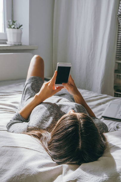 young brunette woman lying on the bed and using smartphone, woman using smartphone at home in the bedroom selective focus - Photo, image