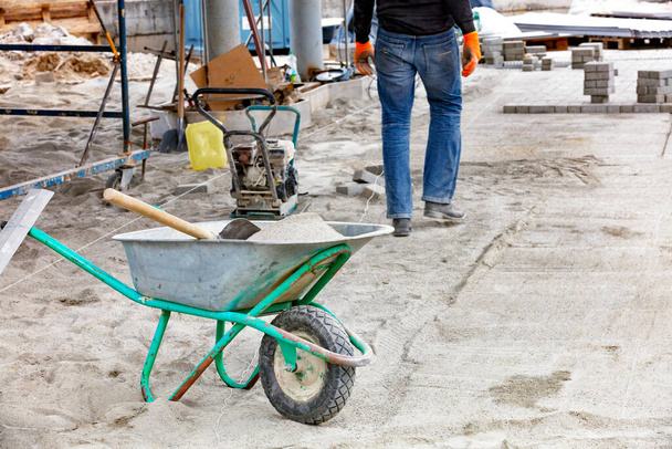 Mit einer Bauschubkarre und einer Schaufel wird auf der Baustelle Sand angeliefert, um Pflasterplatten vor dem Hintergrund des Arbeiters und der Rüttelplatte zu verlegen.. - Foto, Bild