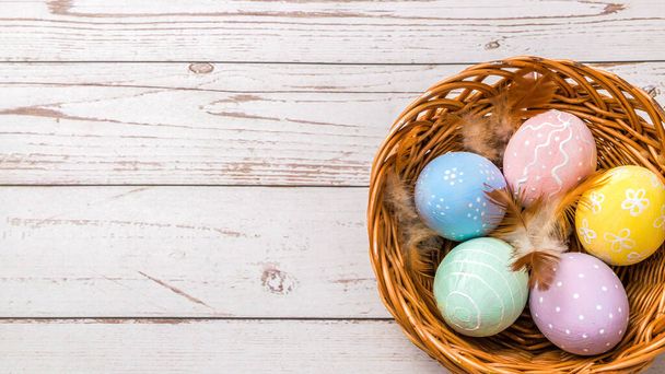 top view of easter eggs hand painted in pastel colors and tender feathers in a wicker basket on wooden table - Φωτογραφία, εικόνα