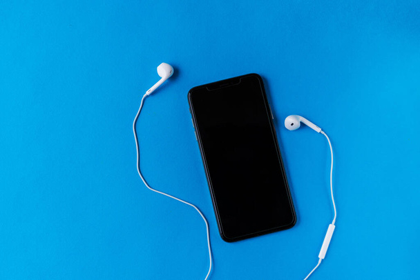 Modern mobiltelefon sötét képernyővel, vezetékes fehér fülhallgatóval körülötte, kék háttérrel. Ötletek posztoláshoz Instagramon vagy Facebookon - Fotó, kép