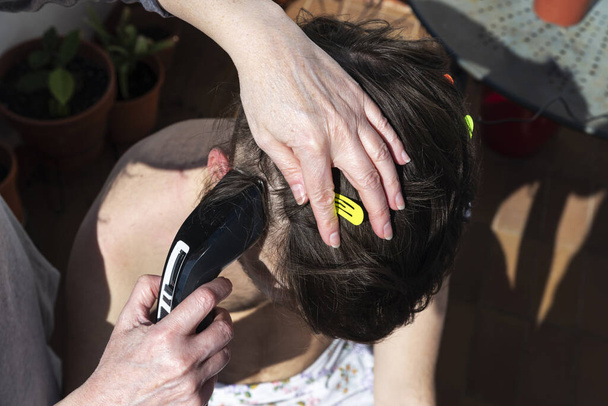 Η μητέρα κόβει τα μαλλιά ενός έφηβου γιου με κουρευτική μηχανή στο μπαλκόνι του σπιτιού του κατά τη διάρκεια της καραντίνας του κορωναϊού στη Βαρκελώνη, Καταλονία, Ισπανία - Φωτογραφία, εικόνα