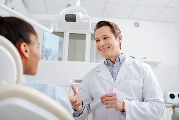 επιλεκτική εστίαση της ευτυχισμένος οδοντίατρος κοιτάζοντας αφροαμερικανός ασθενής και gesturing στην κλινική  - Φωτογραφία, εικόνα