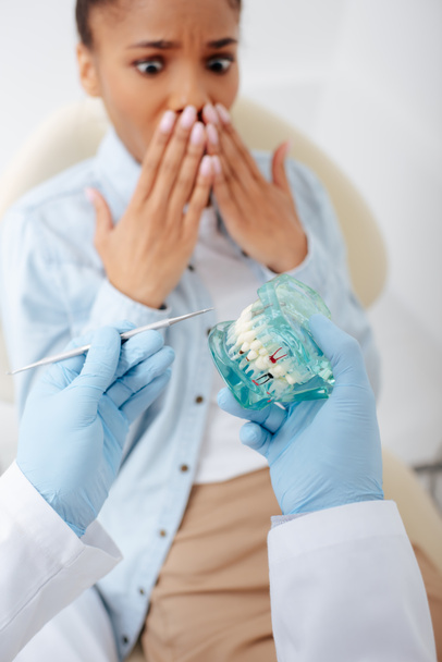 επιλεκτική εστίαση του οδοντίατρου σε γάντια λατέξ κρατώντας τα δόντια μοντέλο και οδοντιατρικό όργανο κοντά σοκαρισμένη αφροαμερικανίδα γυναίκα  - Φωτογραφία, εικόνα
