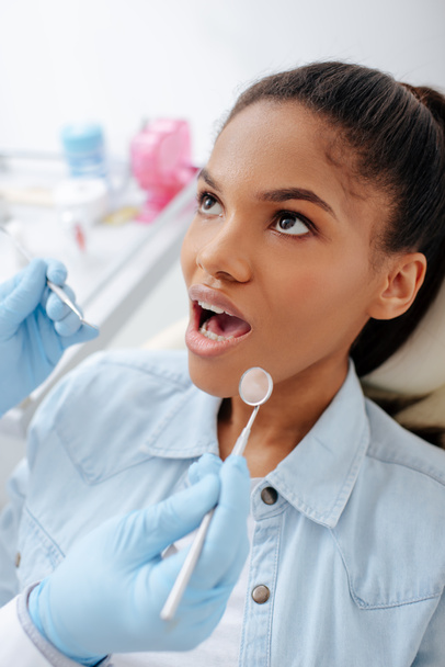 dentista en guantes de látex sosteniendo equipo dental cerca de paciente afroamericano con la boca abierta
  - Foto, imagen