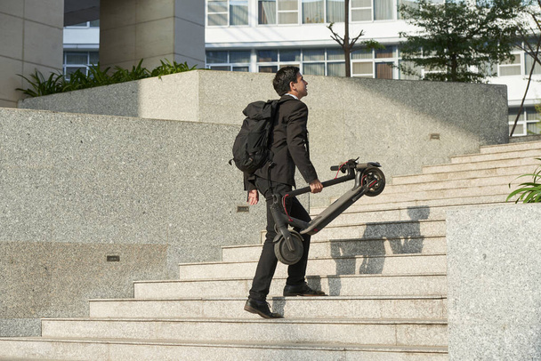 Student walking up the stairs to university with big backpack and electric scooter - 写真・画像