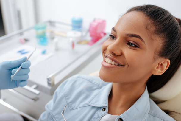orthodontist in het houden van tandheelkundige instrumenten in de buurt gelukkig Afrikaans amerikaanse patiënt in beugels - Foto, afbeelding