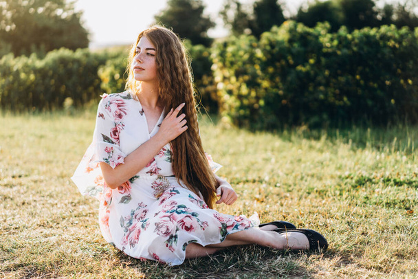 Full length portrait of beautiful woman with long curly hair on currant field background. Girl in a light dress sits on the grass in sunny day. - Foto, Bild