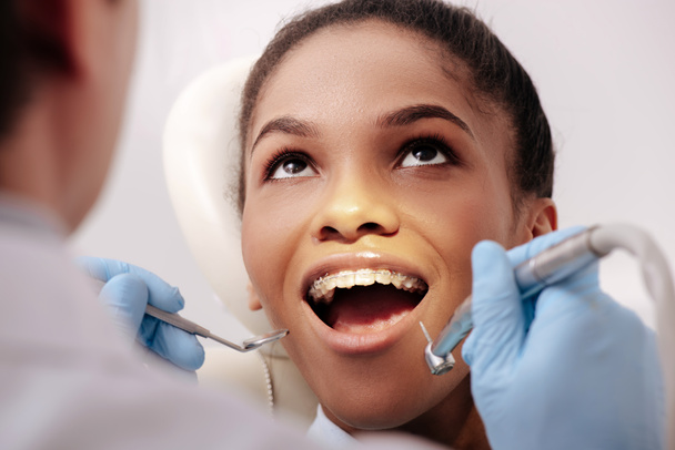 selektywne skupienie się dentysty w lateksowych rękawiczkach trzymającego instrumenty stomatologiczne w pobliżu szczęśliwej, afrykańskiej kobiety z otwartymi ustami  - Zdjęcie, obraz