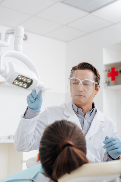 enfoque selectivo del dentista en el escudo facial y guantes de látex tocando lámpara médica cerca del paciente
  - Foto, imagen