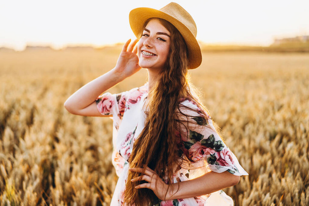 Portrait of a beautiful young woman with curly hair and freckles face. Woman in dress and hat posing in wheat field at sunset and looking at camera. - Foto, Bild