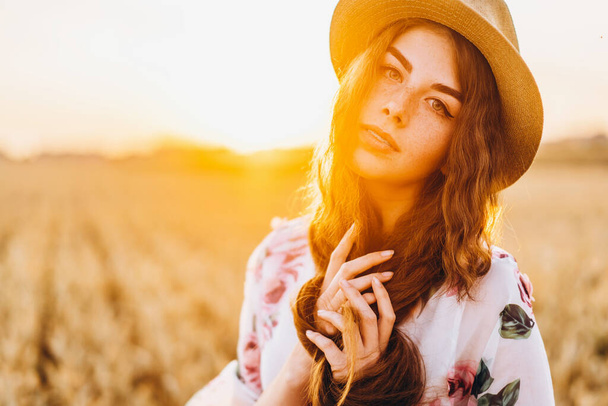 Portret van een mooie jonge vrouw met krullend haar en sproeten gezicht. Vrouw in jurk en hoed poseren in tarweveld bij zonsondergang en kijken naar camera. - Foto, afbeelding