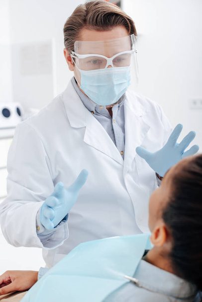 アフリカ系アメリカ人女性に近い顔の盾、医療用マスク、ラテックス手袋の歯科医の選択的焦点  - 写真・画像