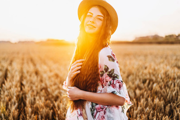 Portrait of a beautiful young woman with curly hair and freckles face. Woman in dress and hat posing in wheat field at sunset and looking at camera. - Фото, изображение