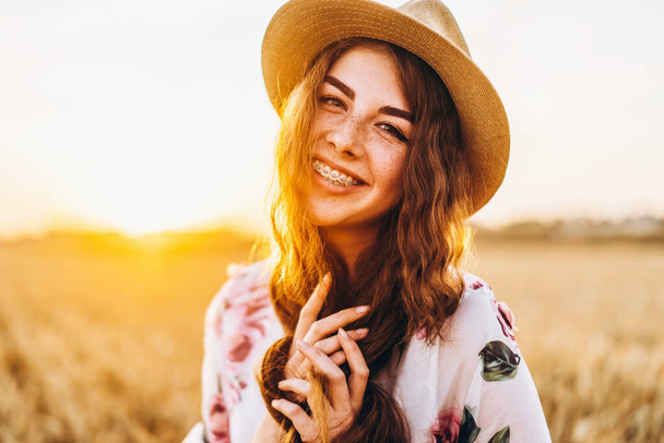 Portrait of a beautiful young woman with curly hair and freckles face. Woman in dress and hat posing in wheat field at sunset and looking at camera. - Fotografie, Obrázek