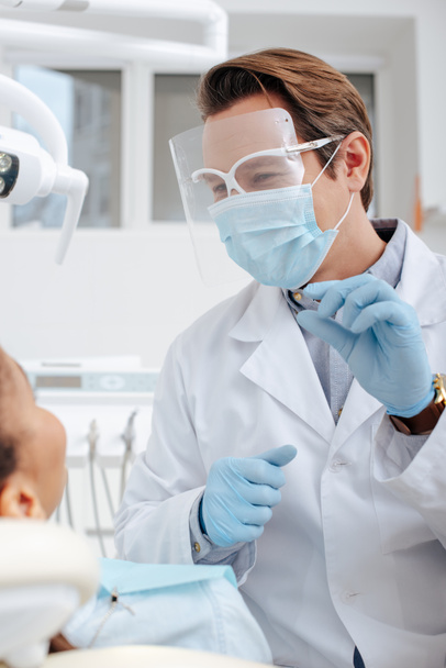 селективный фокус стоматолога в медицинской маске, защитных и латексных перчатках, держащих стоматологический инструмент рядом с африканской американкой
  - Фото, изображение