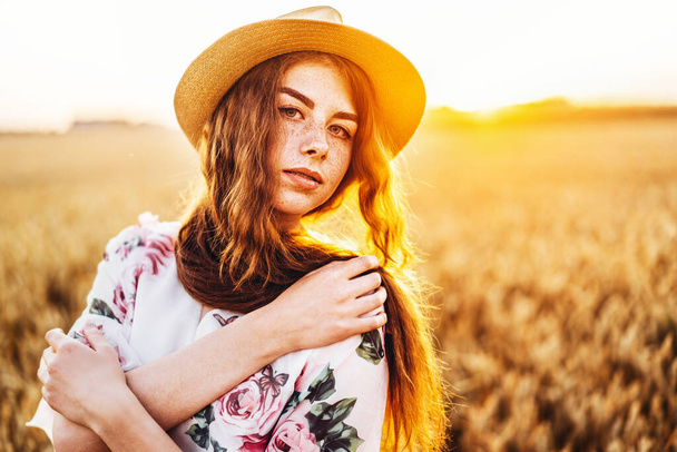 Portrait of a beautiful young woman with curly hair and freckles face. Woman in dress and hat posing in wheat field at sunset and looking at camera. - 写真・画像