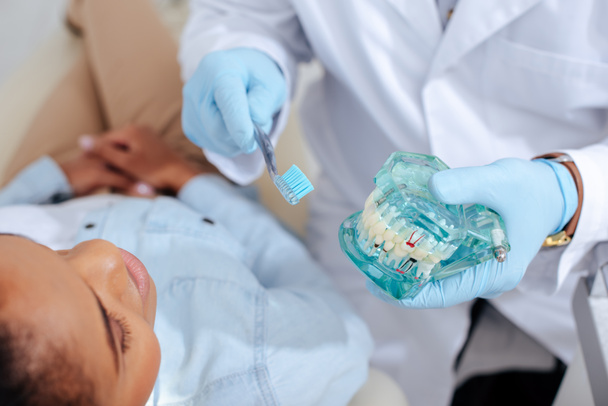 enfoque selectivo del dentista en guantes de látex que sostienen el modelo de dientes y el cepillo de dientes cerca del paciente afroamericano
  - Foto, Imagen