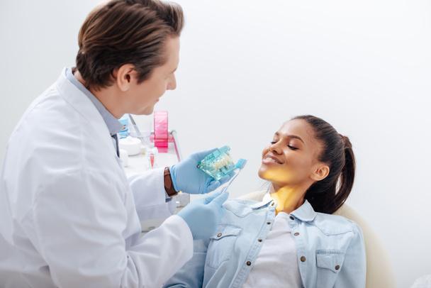 селективний фокус стоматолога в латексних рукавичках, що тримає модель зубів і зубну щітку біля щасливого афроамериканського пацієнта
  - Фото, зображення